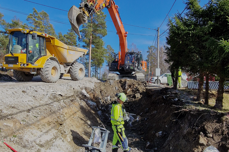 Person som jobbar med att lägga ner ledningar i marken, i bakgrunden syns en dumper och grävmaskin.